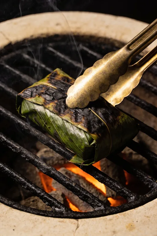 Tungku — charred banana-leaf parcel on a glowing charcoal grill, brass tongs entering the frame